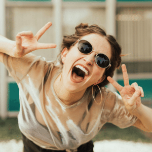 Frau mit Sonnenbrille, lachend, mit beiden Händen V-Zeichen
