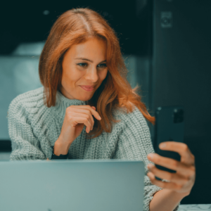 Frau vor Laptop blickt zufrieden auf ihr Handy